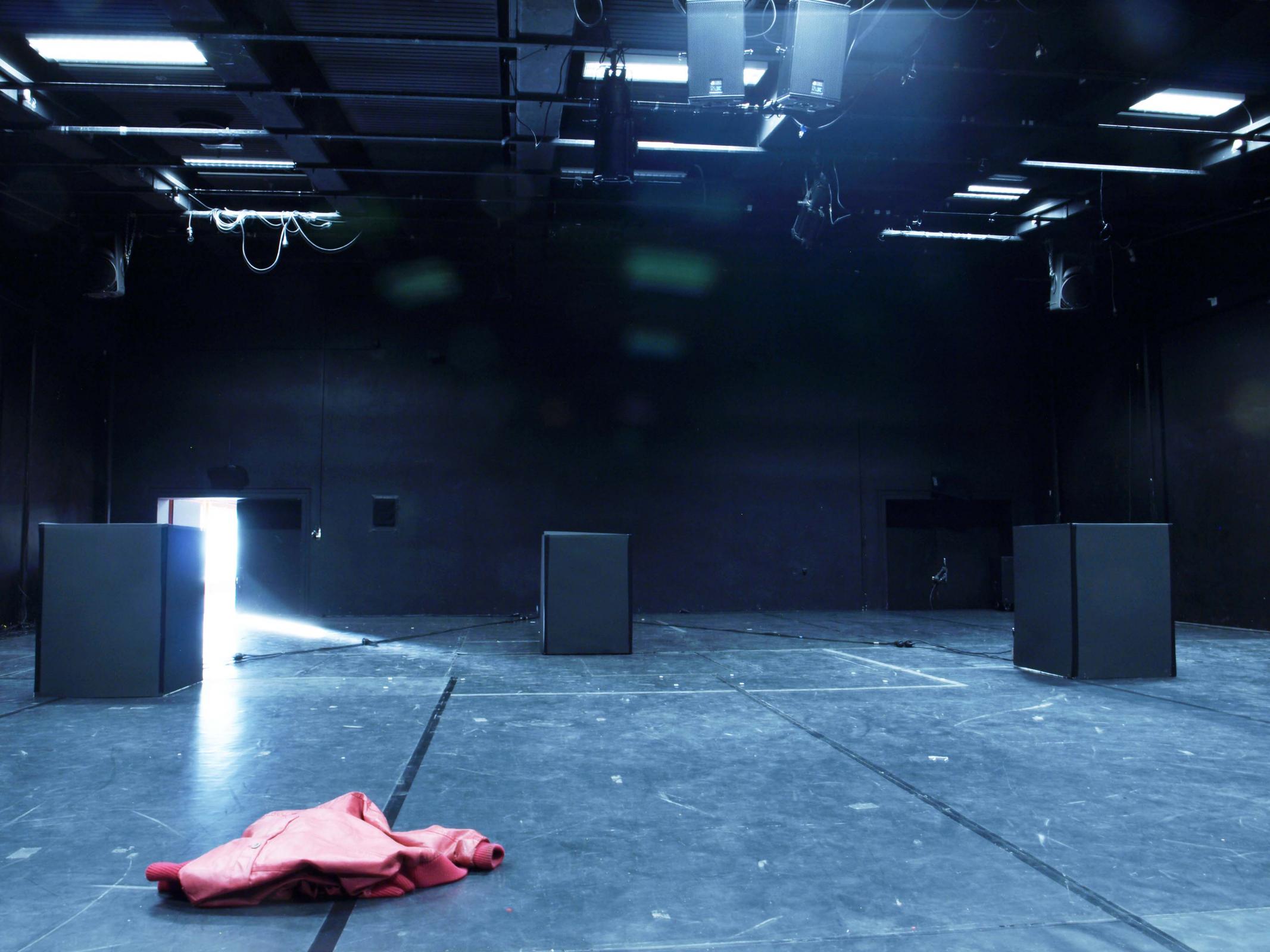 Auf einer Bühne stehen drei schwarze Kuben am Bildrand und in der Mitte. Eine rote Jacke liegt im Vordergrund